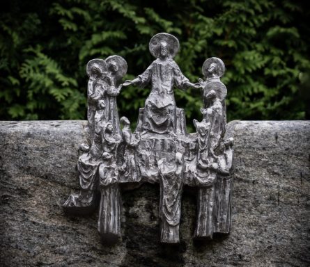 Detailaufnahme einer religiösen Skulptur auf einem Grabstein, umgeben von grünen Lebensbäumen.