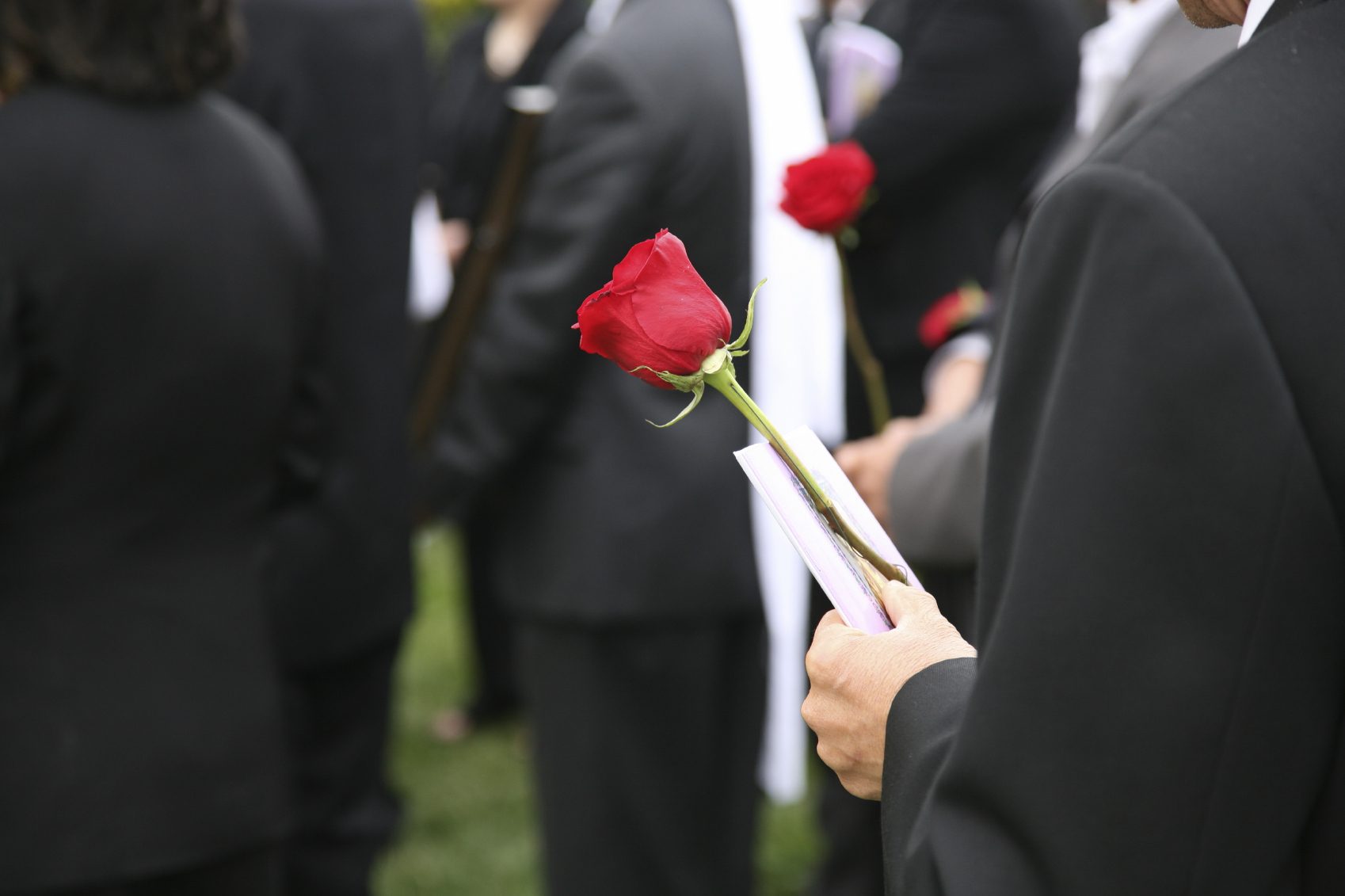 Trauergäste - Mehrere Personen halten rote Rosen in der Hand  bei einer Trauerfeier während einer einfühlsamen Abschiedsfotografie