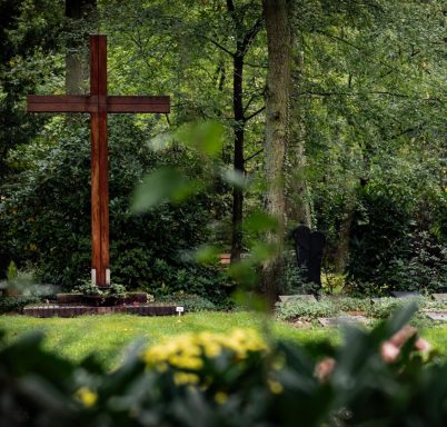 Foto eines großen Holzkreuzes auf einem Friedhof, umgeben von Bäumen und grüner Wiese.